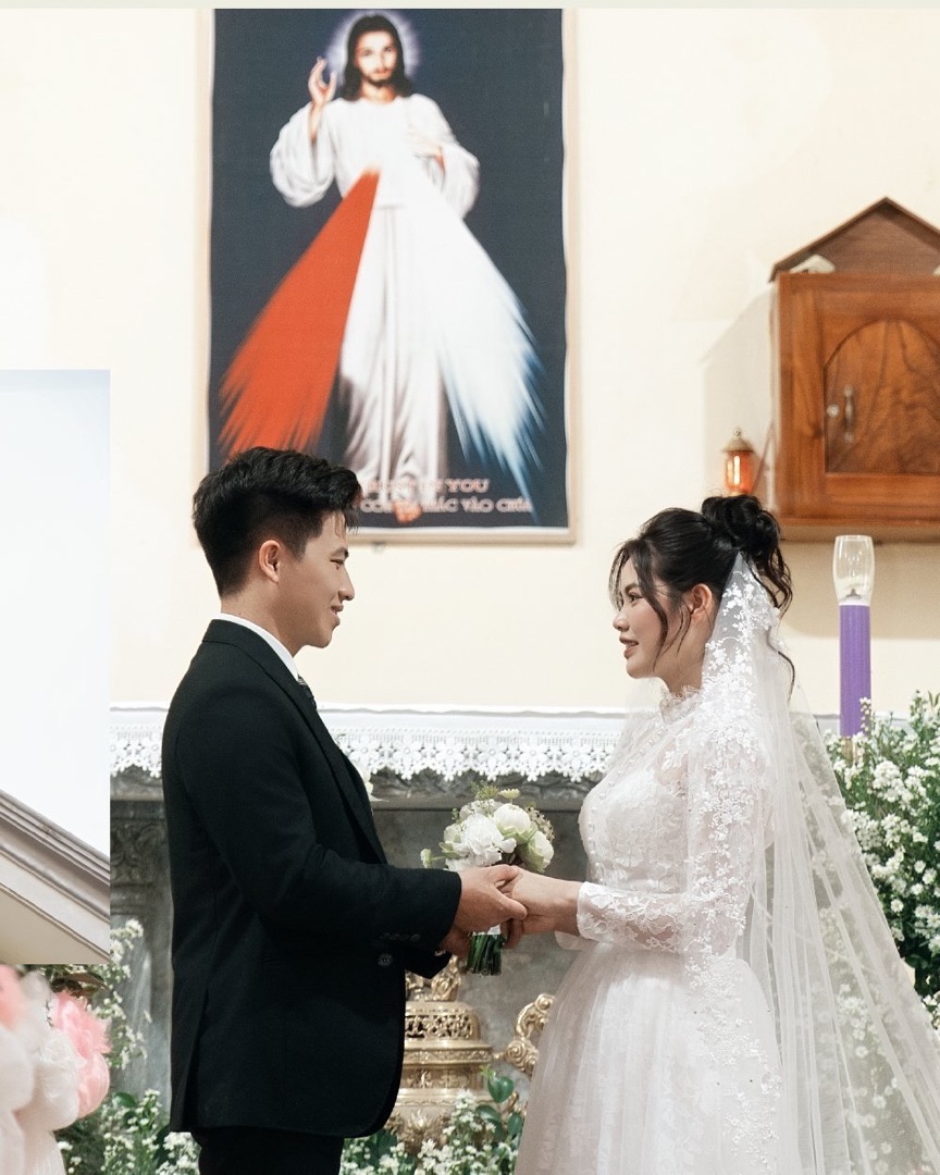 Cô dâu chú rể nắm tay nhau nhìn nhau trước ảnh Lòng Chúa Thương Xót trong nhà thờ