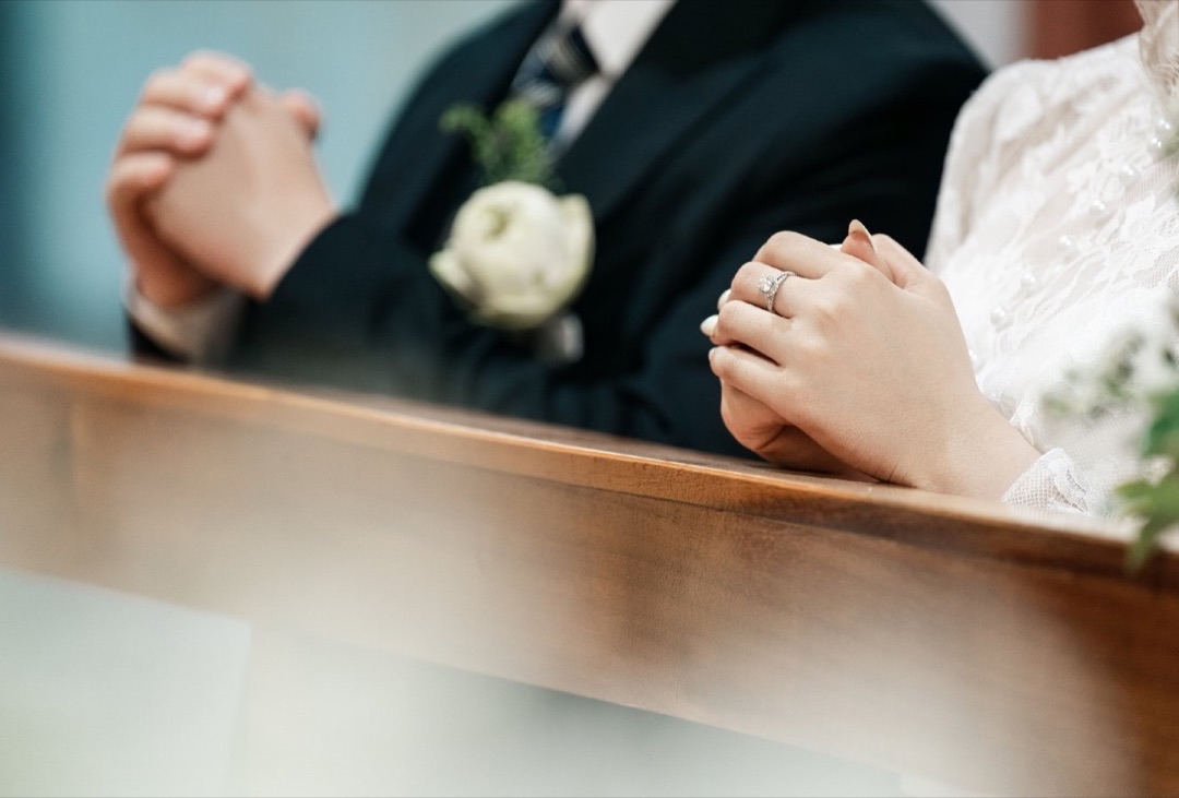 Cận cảnh đôi bàn tay cô dâu và chú rể đan lại cầu nguyện, tựa trên ghế quỳ gỗ trong nhà thờ